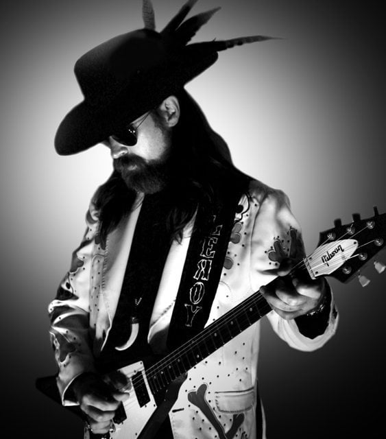 Black and white portrait of Leroy Powell playing a white Flying V guitar in a decorated stage jacket and hat