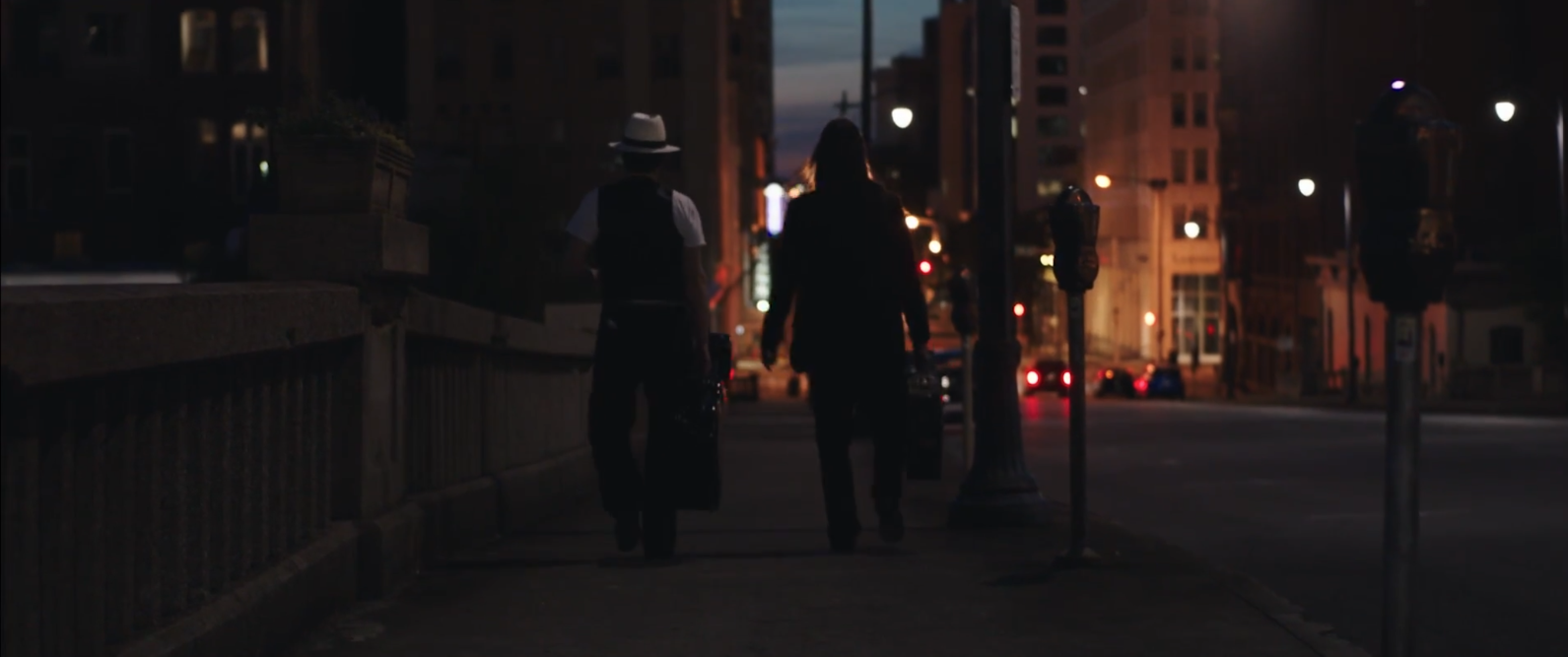 Two musicians carrying guitar cases down a city sidewalk at night